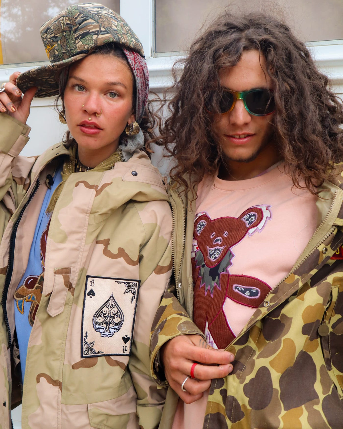 Close-up of two people in desert camo jacket with ace of spades patch and pink bear graphic tee