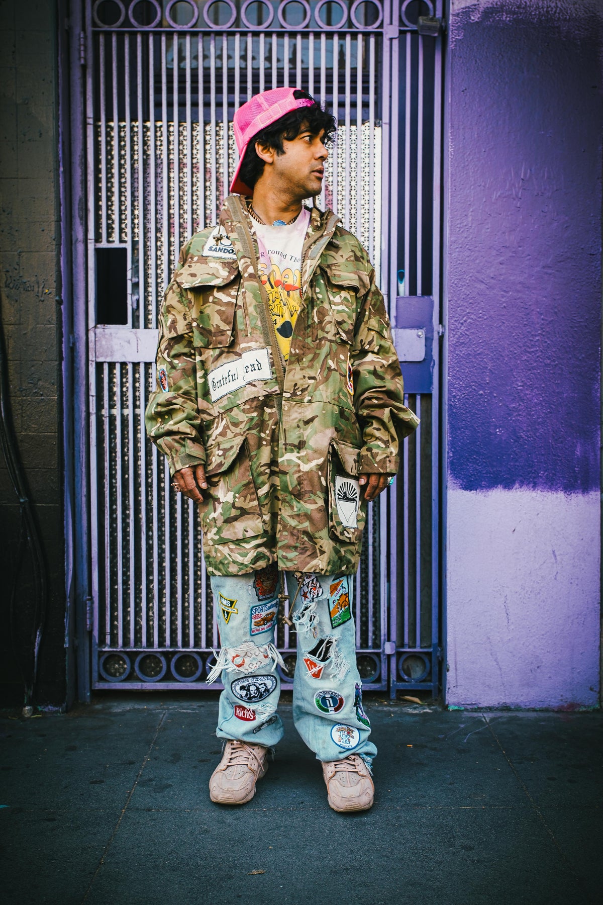 Man in camo jacket and pink cap with patched jeans posing against a purple wall