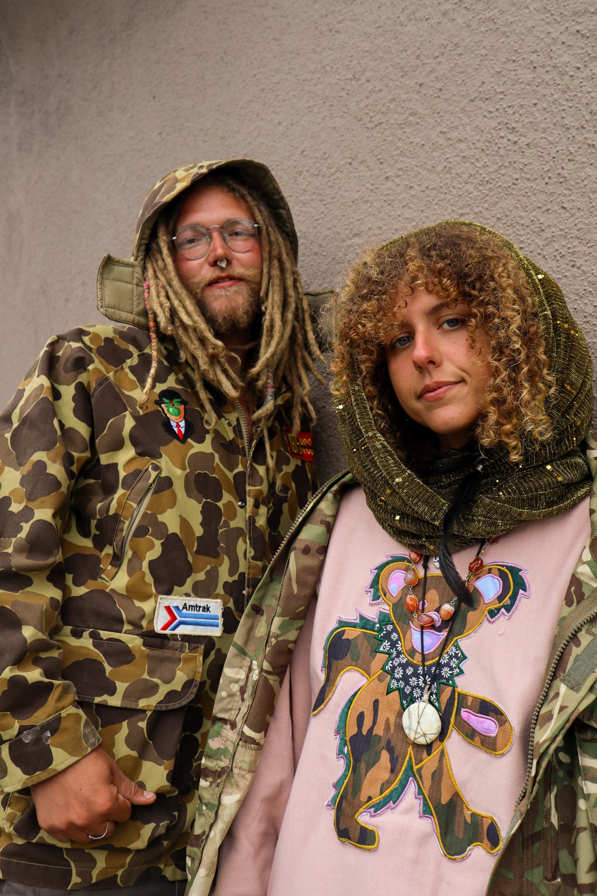 Couple in layered camo and pink bear graphic hoodie leaning against stucco wall