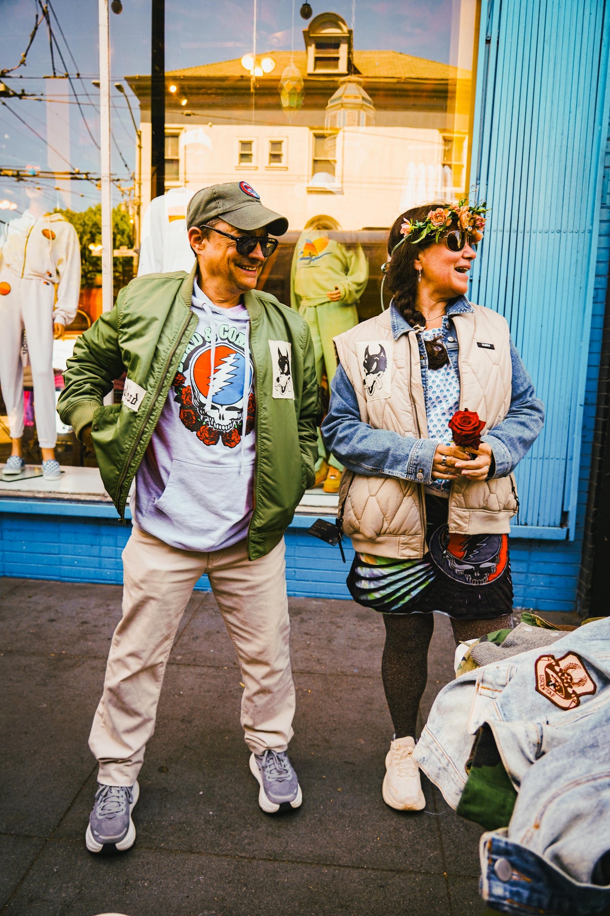 Couple laughing outside a blue storefront in green bomber with Grateful Dead tee and denim flower crown