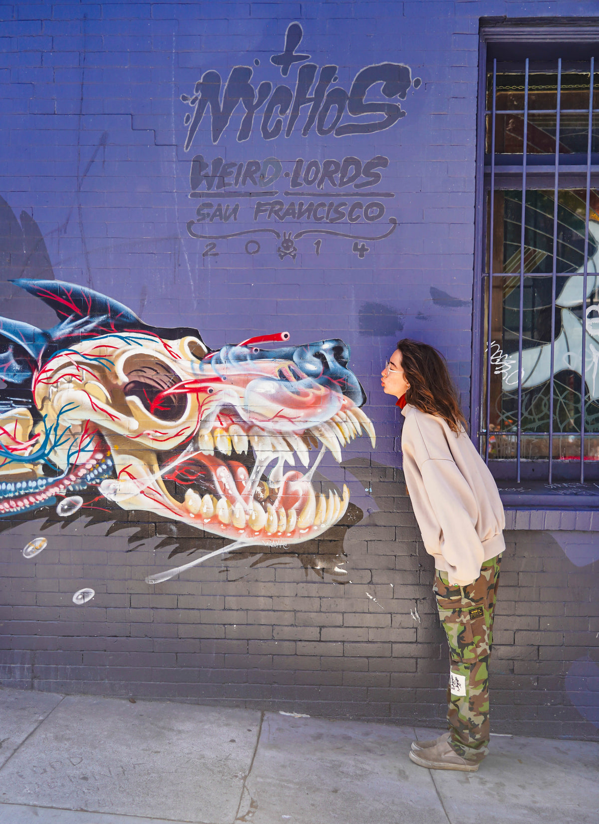 Woman standing in front of Nychos mural in San Francisco wearing beige hoodie and camo pants