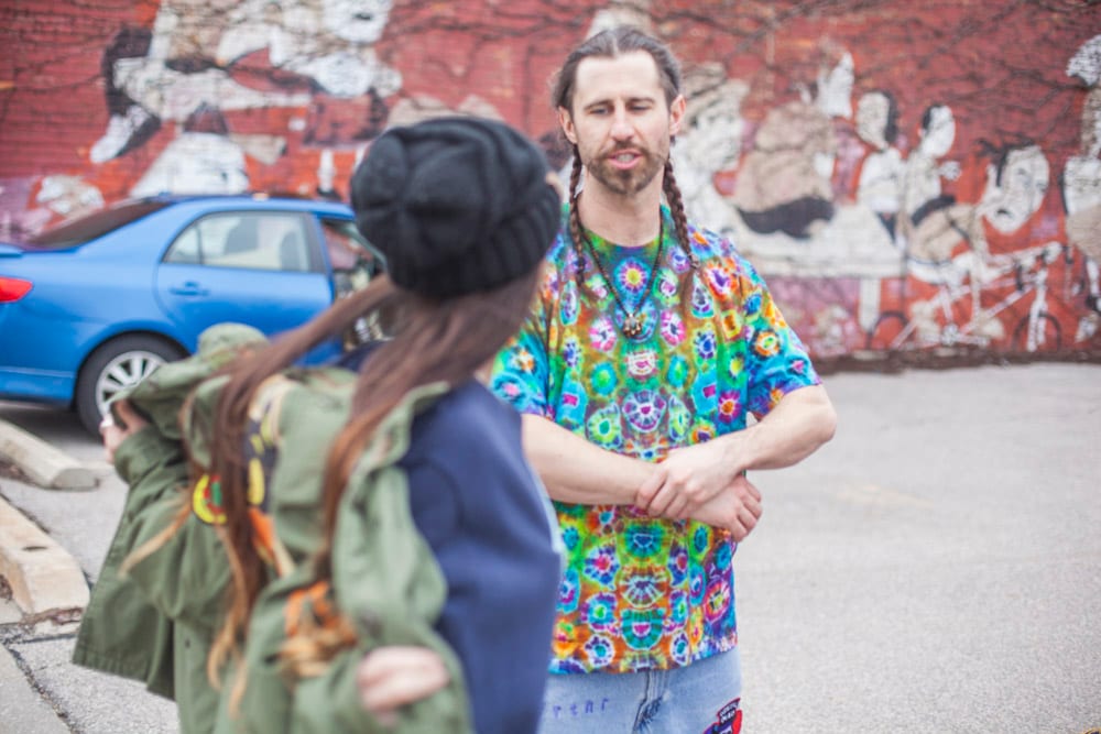 Man in colorful tie-dye shirt with braids talking to woman in army jacket by a street mural