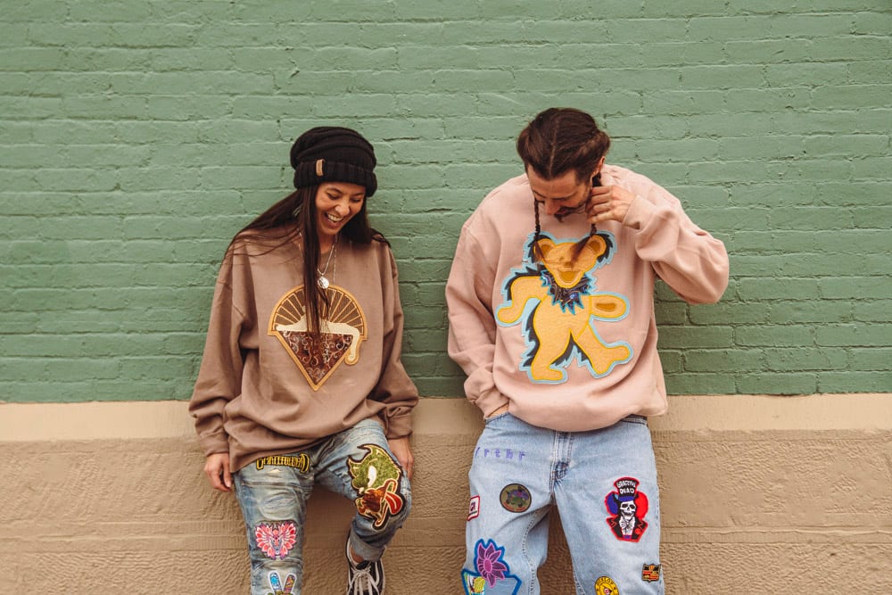Two people laughing against green brick wall in graphic sweatshirts and patched denim