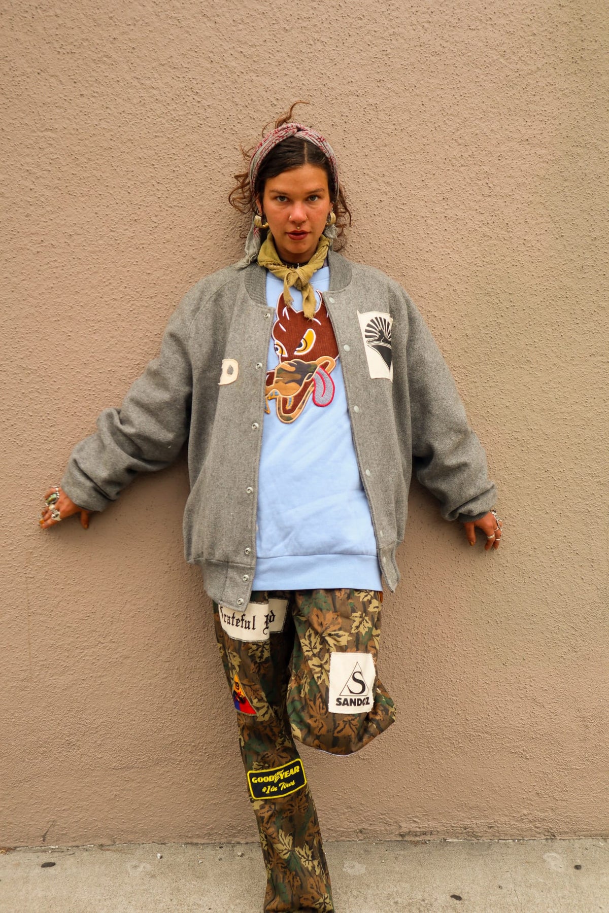 Woman in gray varsity jacket with dragon graphic and camo pants leaning against a wall