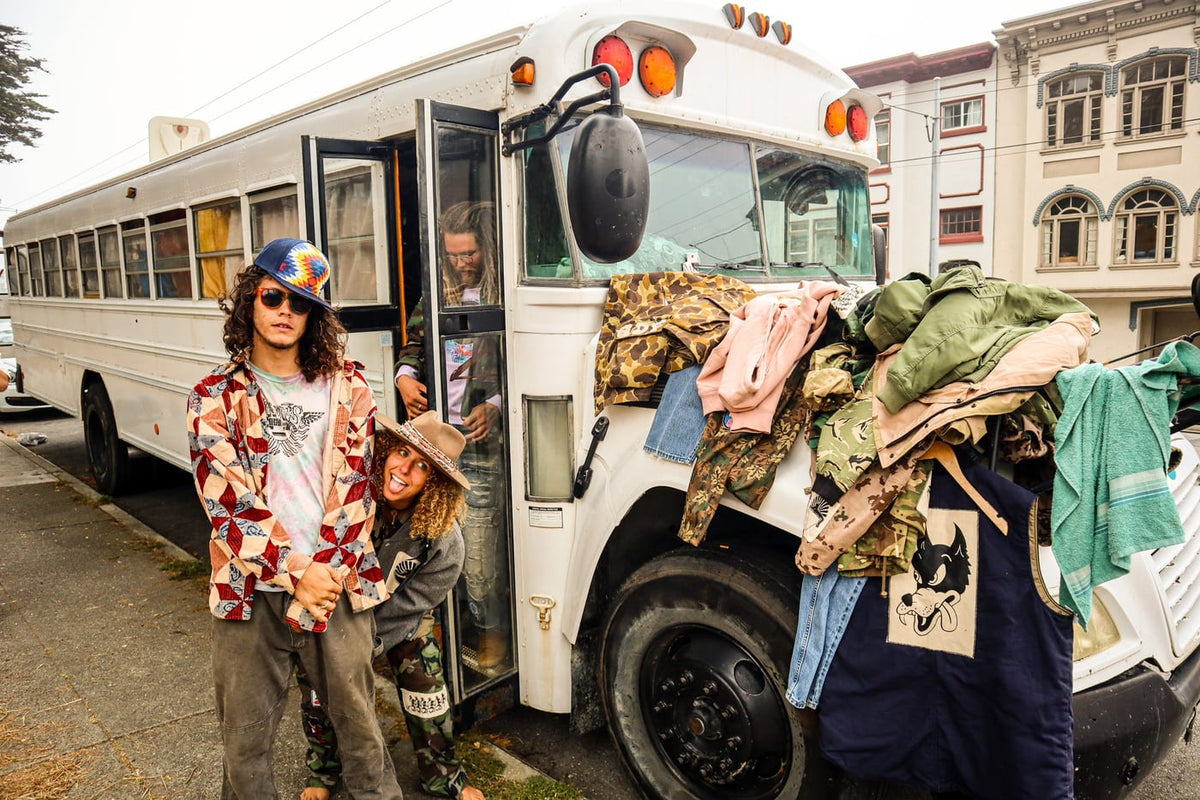 Two people posing by a white bus with a clothing rack at a street pop-up